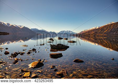 Rocky Shore Of Lake Ohau With The Snow Capped Southern Alps Reflected In The Calm Water