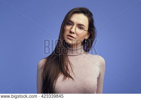 Girl Looks Seriously At Camera, Wears Round Spectacles Against Violet Wall