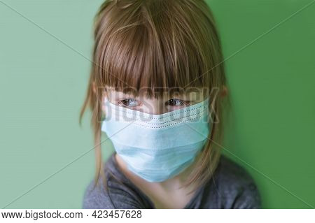A Sad Little Girl In A Medical Mask On A Green Background. Disposable Medical Mask On The Child's Fa