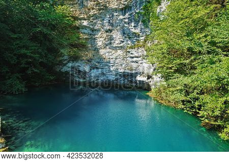 The Alpine Lake Is Surrounded By Sheer Cliffs With Green Vegetation. Reflection And Shadows On Calm 