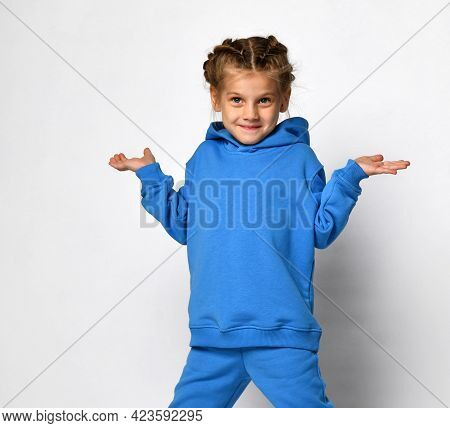 Little Sly Girl In A Blue Sports Suit Shrugs While Standing On A White Background. Stylish Child Wit