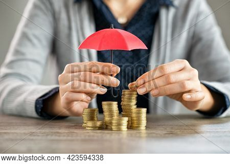Young woman hands holding red umbrella over stacked coin on table. Female hand holding a small umbrella to protect heaps of coins while saving them. Financial security and savings protection concept.