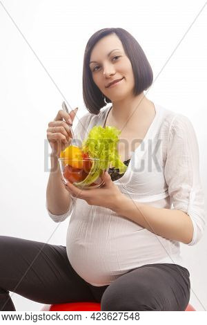 Young Pregnant Brunette Woman Sitting On Fitball And Eating Fresh Vegetables In Glass Bowl Over Whit