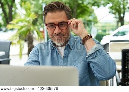 Outdoor portrait of mid adult man in 50s, happy confident smile. Working with laptop computer. Mature age, middle age, bearded, glasses.