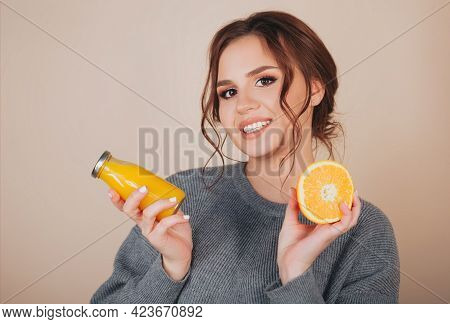 Brunette Female Wearing Grey Wooven Sweater Holding Orange Slice And Bottele With Orange Juice In He