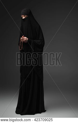 Full Length View Of Muslim Woman In Black Niqab Praying With Rosary On Dark Grey Background