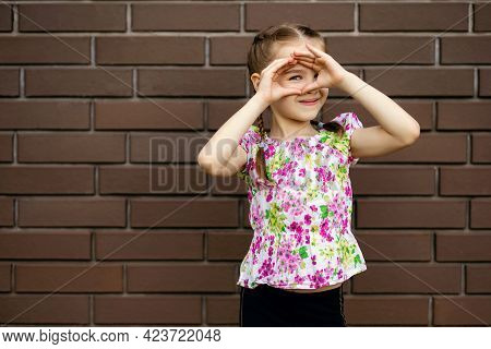 Cheerful Little Girl Child Spies Closing Her Eyes With Her Hands. A Cute Girl In Bright Clothes Stan