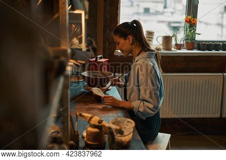 Friendly Brunette Ceramicist During Painting Process Of Clay Tableware In Pottery Workshop