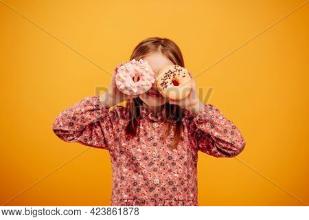 Little girl playing and holding donuts as glasses isolated on yellow background with copyspace. Pretty female kid with sweets