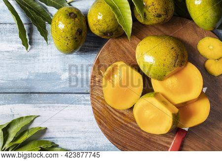 Top View Of Ripe Mango Slices On Wooden Chopping Board Along With Mangoes And Leaves On Wooden Table