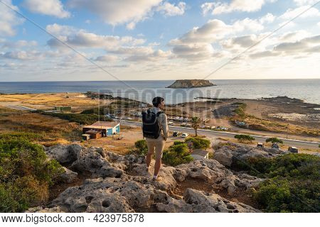 Man With Backpack Is Hiking Along A Rocky Coast At Sunset Near The Mediterranean Sea In Cyprus. Male