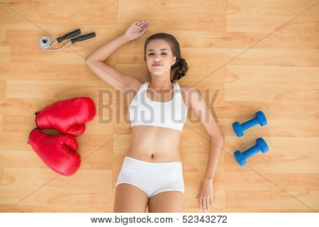 Content sporty brunette lying next to jump rope, red boxing gloves and dumbbells on the floor