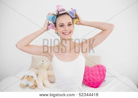 Pleased natural brown haired woman fixing her hair curlers in bright bedroom