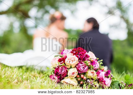 Wedding couple of bride and groom sitting on the meadow with bridal bouquet behind them, candid shot