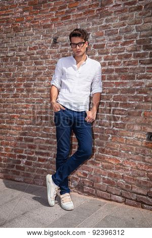 Attractive young man posnig with his hands in pockets while leaning on a brick wall/