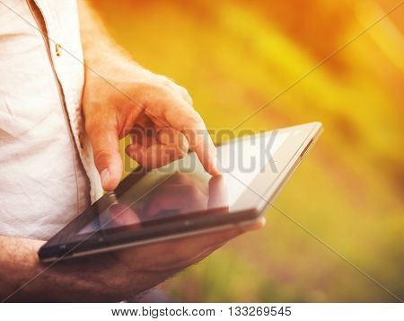 Smart casual man with digital tablet outdoors, close up. Focus on finger. Sunkissed effect with sunflares. Modern remote work concept.