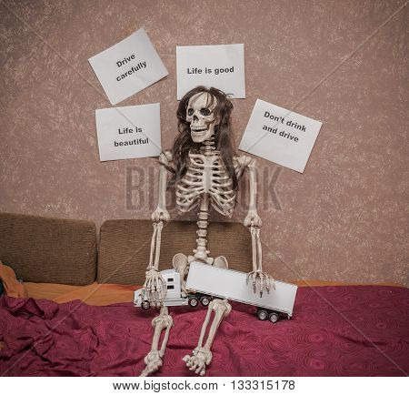 Skeleton in a wig sitting on the bed with truck and trailer model on his legs, leaning against the wall with various messages about safety driving behavior and consequences on the road