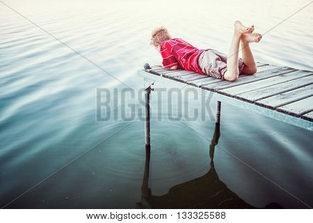 Boy laying on a dock by a lake, vintage toned image.