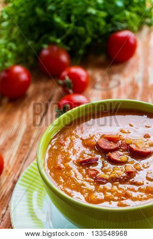 Soup: tomato soup with smoked sausage, tomatoes and lentil. Green bowl on wooden table. Small cherry tomatoes and green lettuce on table.