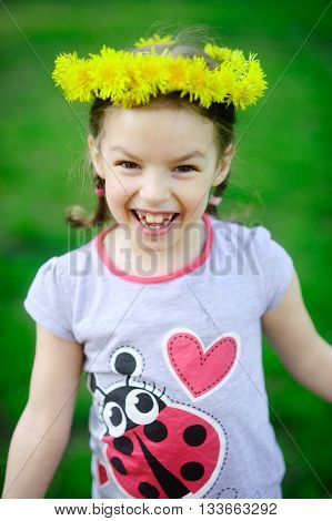 The little girl in a wreath from yellow dandelions. The cheerful girl on a green background. Hair are braided in small braids. On the head a beautiful wreath. The wide smile bares large teeth.