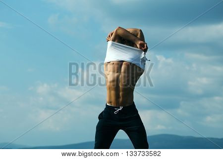 young macho man model athlete with muscular sexy body and wet bare chest outdoor on sky background undressing white shirt