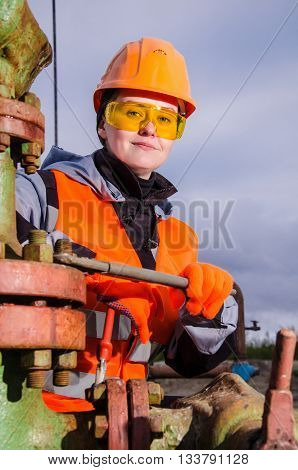 Woman engineer in the oil field repairing wellhead with the wrench wearing orange helmet and work clothes. Oil and gas concept.