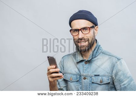Stusio Shot Of Happy Bearded Man With Stubble Holds Modern Smart Phone In Hands, Enjoys Online Commu