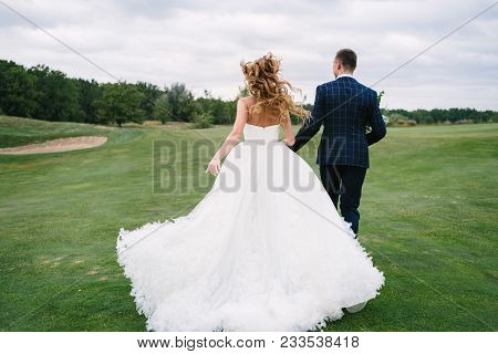 Full Length Body Portrait Of Young Bride And Groom Running On Green Grass Of Golf Course, Back View.
