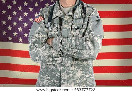 Closeup of a military doctor with a stethoscope around his neck in front of American Flag. The man is wearing camouflage fatigues also called ACU and has his arms crossed.