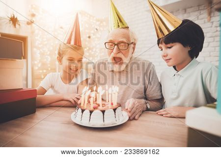 Grandfather, Grandson And Granddaughter At Table At Home. Grandpa Is Blowing Candles On Birthday Cak