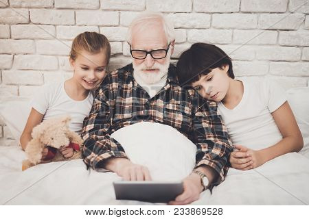 Grandfather, Grandson And Granddaughter At Home. Grandpa And Children Are Using Tablet In Bed.