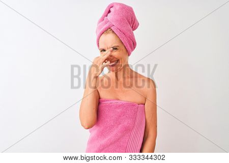Middle age woman wearing shower towel after bath standing over isolated white background smelling something stinky and disgusting, intolerable smell, holding breath with fingers on nose. Bad smells