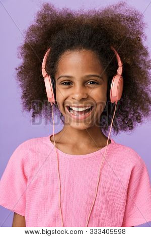 Image of a happy smiling optimistic young african girl kid posing isolated over purple wall background listening music with headphones.