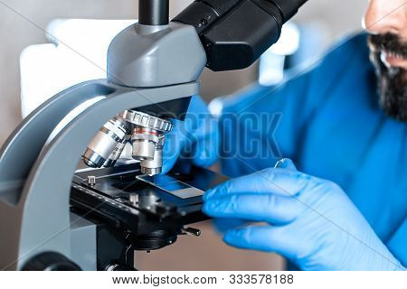 Male Laboratory Assistant Examining Biomaterial Samples In A Microscope. Cllose Up Hands In Blue Rub