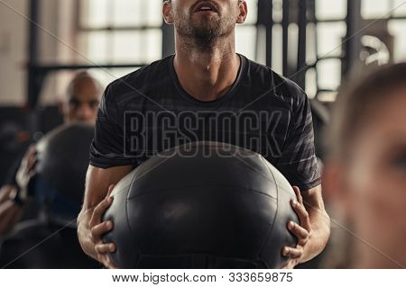 Closeup of sportsman doing weight lifting with heavy medicine ball. Determined man doing training in fitness center. Young man doing intensive squat workout at gym with fitness ball.