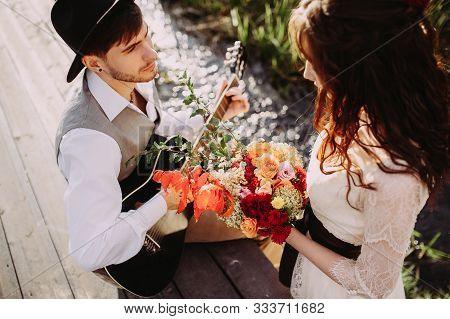 Lovestory On The Background Of A Mountain River. Stylish Guy In A Hat Plays The Guitar. Beautiful Gi