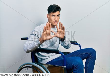 Young hispanic man sitting on wheelchair moving away hands palms showing refusal and denial with afraid and disgusting expression. stop and forbidden. 