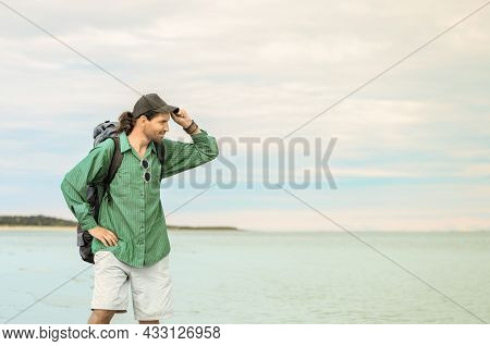 Traveler With His Tourist Backpack Is Standing Against A Sea Background. Man Is Taking Off His Baseb