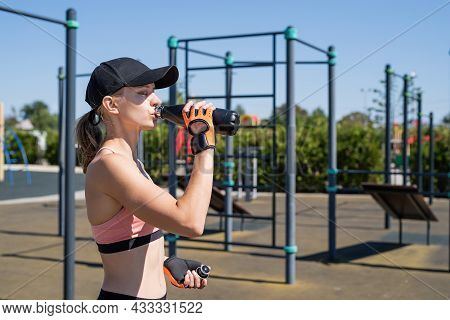 Young Sportive Woman In Sport Gloves Drinking Water On Sport Ground
