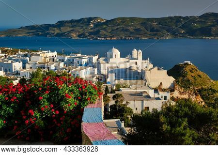 Picturesque scenic view of Greek town Plaka on Milos island over red geranium flowers and Orthodox greek church. Plaka village, Milos island, Greece. Focus on buildings