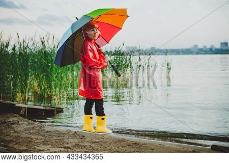 Boy In A Red Raincoat And Yellow Rubber Boots Stands At River Bank And Holding Rainbow Umbrella. Sch