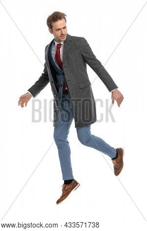 cool dynamic young man in blue suit with grey jacket looking to side and jumping in the air on white background in studio, full body