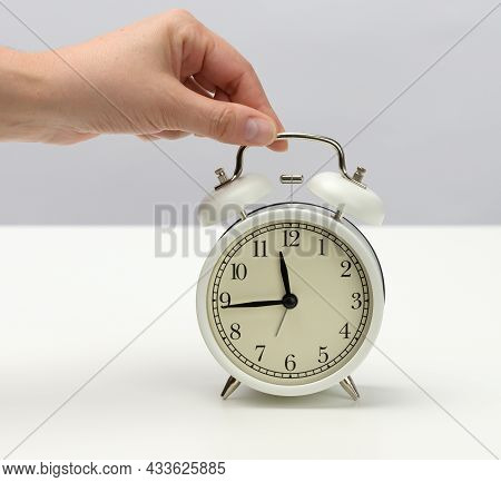 Female Hand Holds A White Metal Alarm Clock On A White Table, The Time Is Fifteen To Twelve. Concept