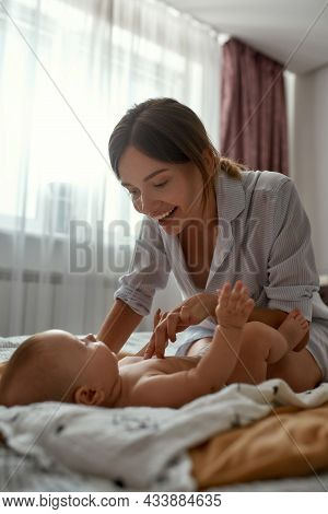 Portrait Of Baby And Mom In Modern Bedroom Interior. Fun-loving Mother Playing With Adorable Naked B