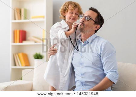 It is very important. Charming little boy in a hospital gown examining a cheek of his father who making a grimace