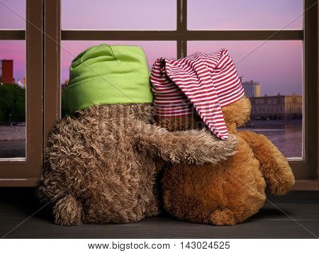 Two friends or fans to watch the sunset in the window. Toys colorful hats bear cubs. Embrace the window. Concept - love friendship support