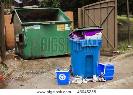 An overflowing recycling bin sits next to trash dumpster.