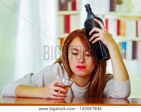 Woman wearing white sweater sitting by bar counter lying over desk holding glass and bottle to head, drunk depressed facial expression, alcoholic concept.