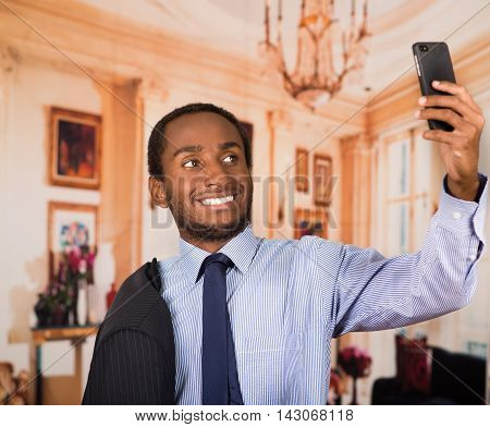Handsome man wearing business suit taking a selfie with mobile phone, as seen from front angle, lobbby background.