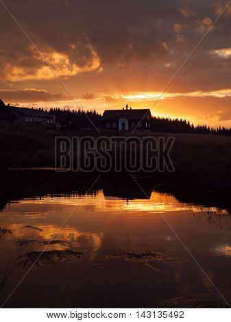 Mountain sunset with river reflection and dark silhouette of forest and rooftop, Jizerka village, Jizera Mountains, Czech Republic